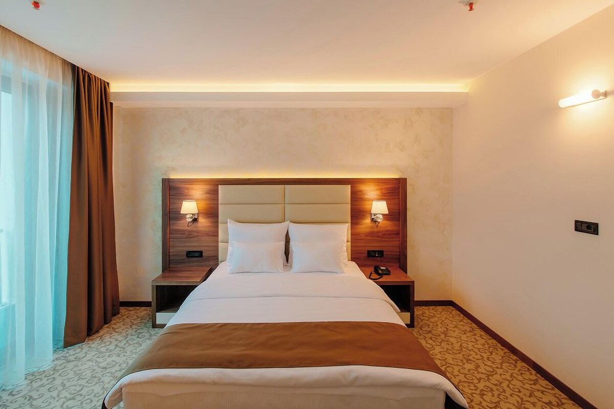 A hotel room with a large bed, two bedside lamps, a nightstand, and a window with curtains.