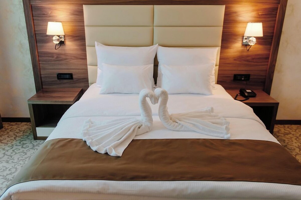A hotel room with a bed, two bedside lamps, and a heart-shaped towel decoration.