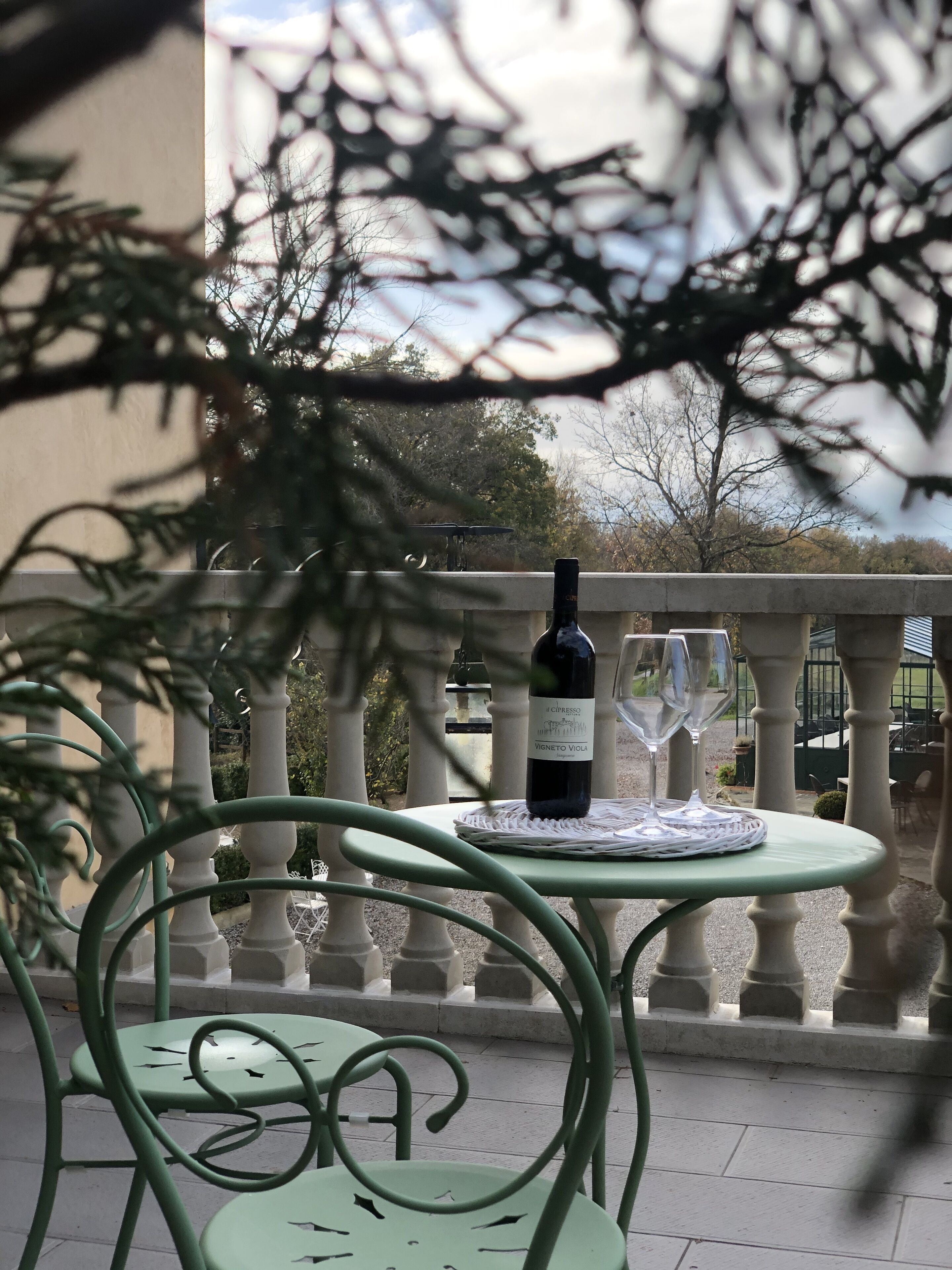 Une table en métal vert, sur laquelle sont posés une bouteille de vin et un verre, le tout placé sur une nappe ronde, avec en arrière-plan une balustrade blanche.
