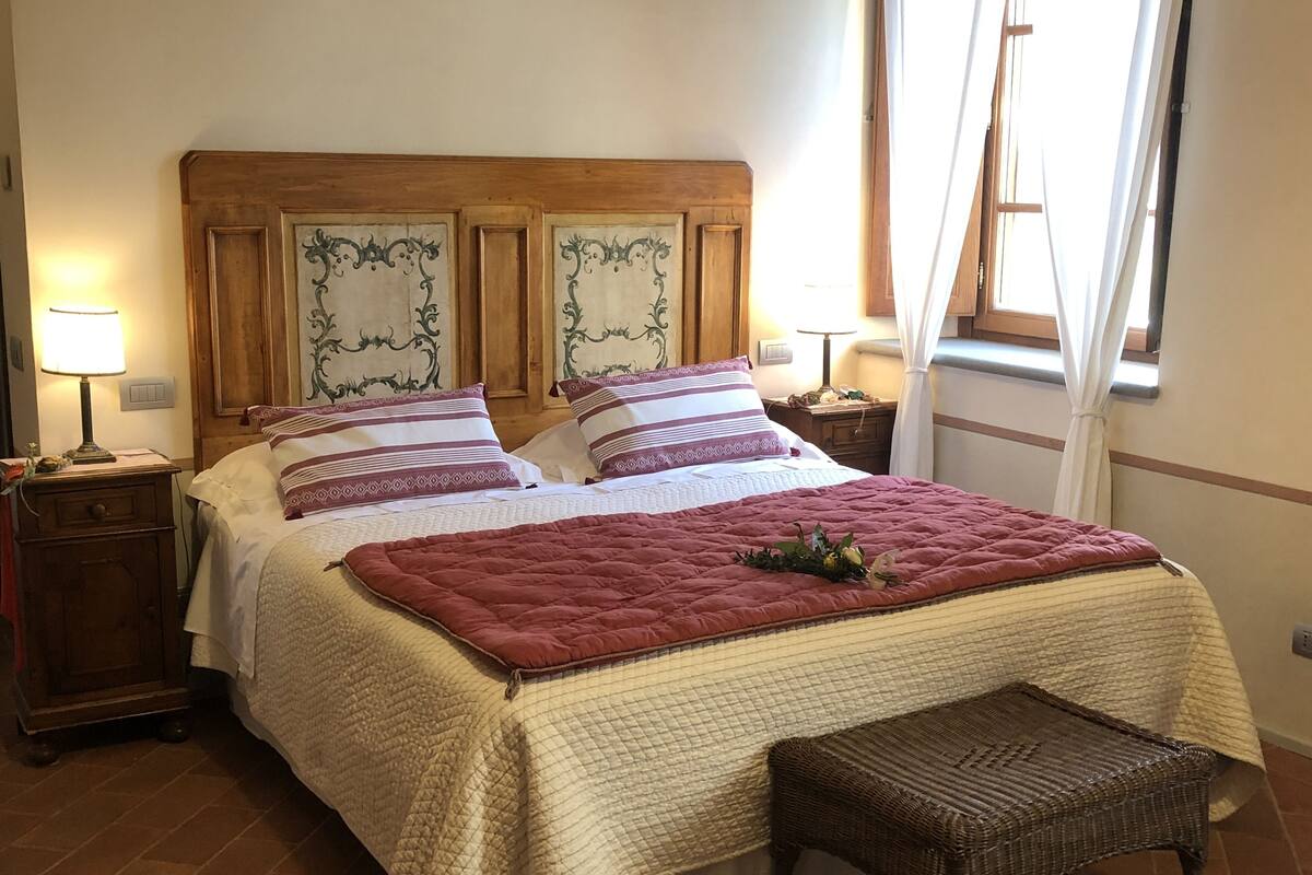 A neatly made bed with a quilted headboard, two bedside tables with lamps, a wicker bench, and a window with curtains.