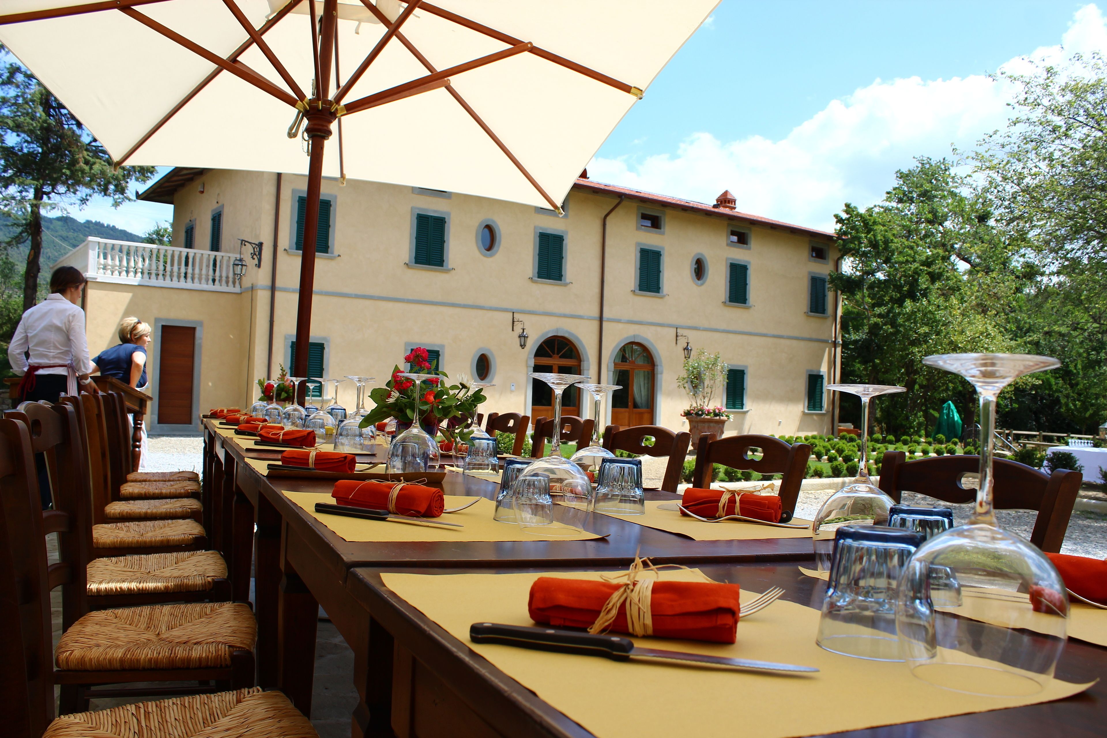 A dining area with tables set for two, outdoor umbrellas, and a building with a balcony in the background.