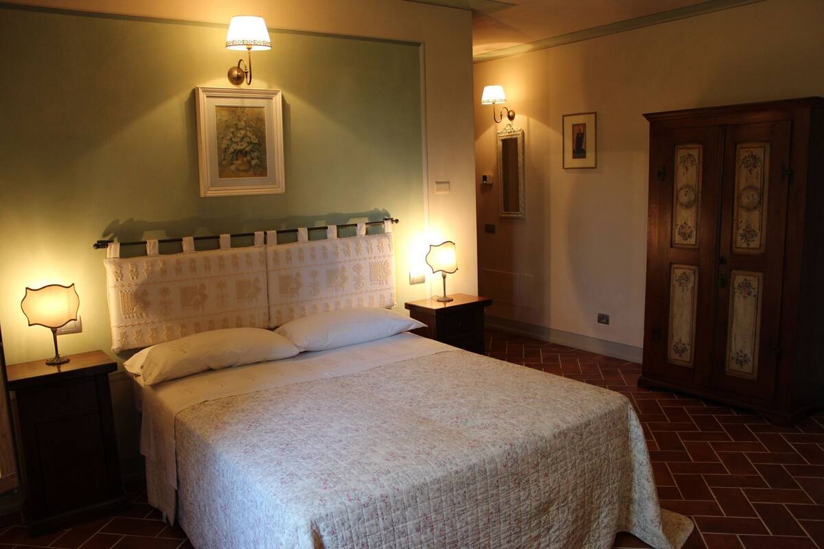 A neatly made bed with a floral bedspread, two bedside lamps, a framed picture above the bed, and a wooden wardrobe with floral designs.