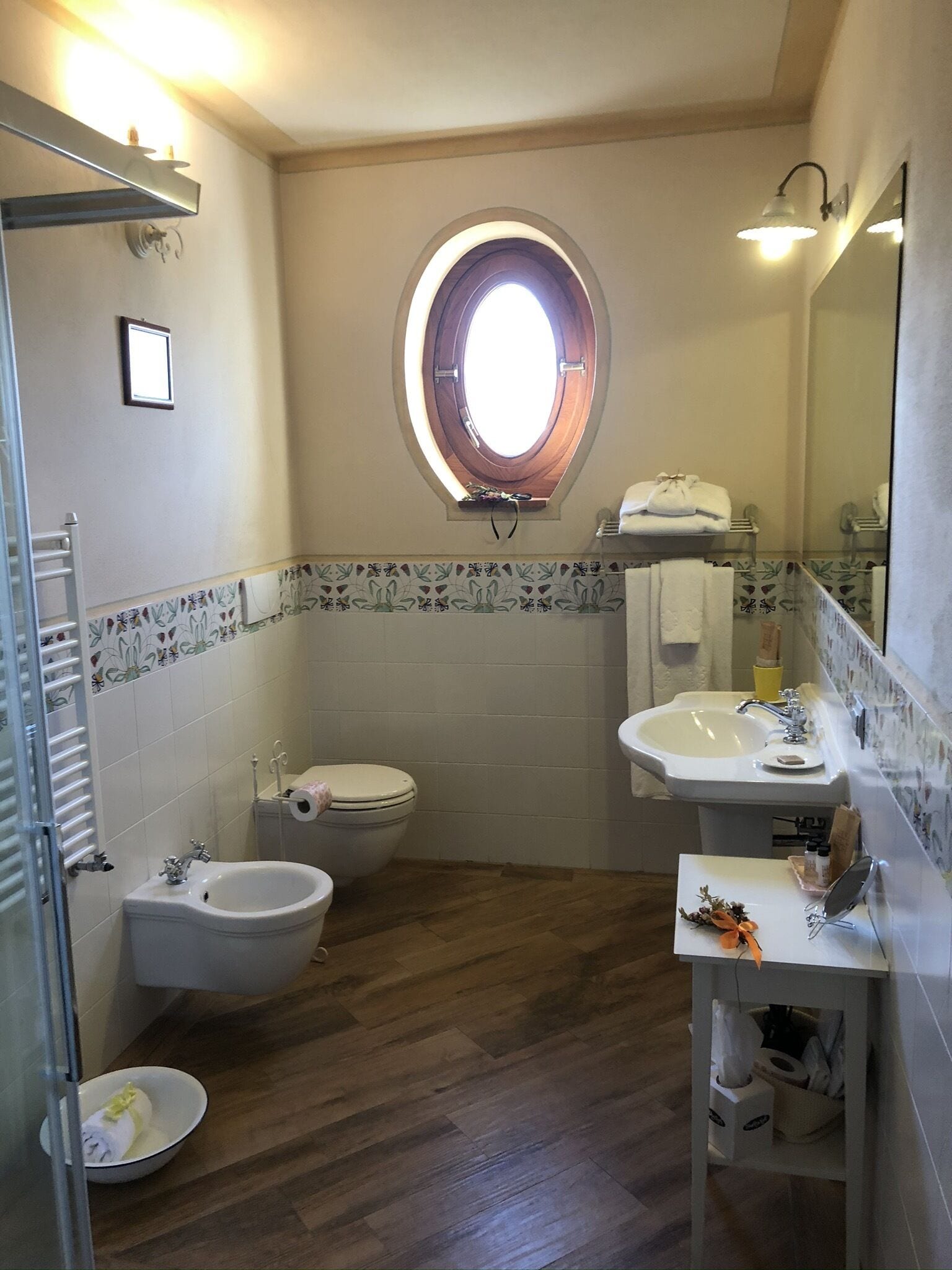 A bathroom with a wooden floor, a round skylight, a bidet, a toilet, a sink, and a mirror.