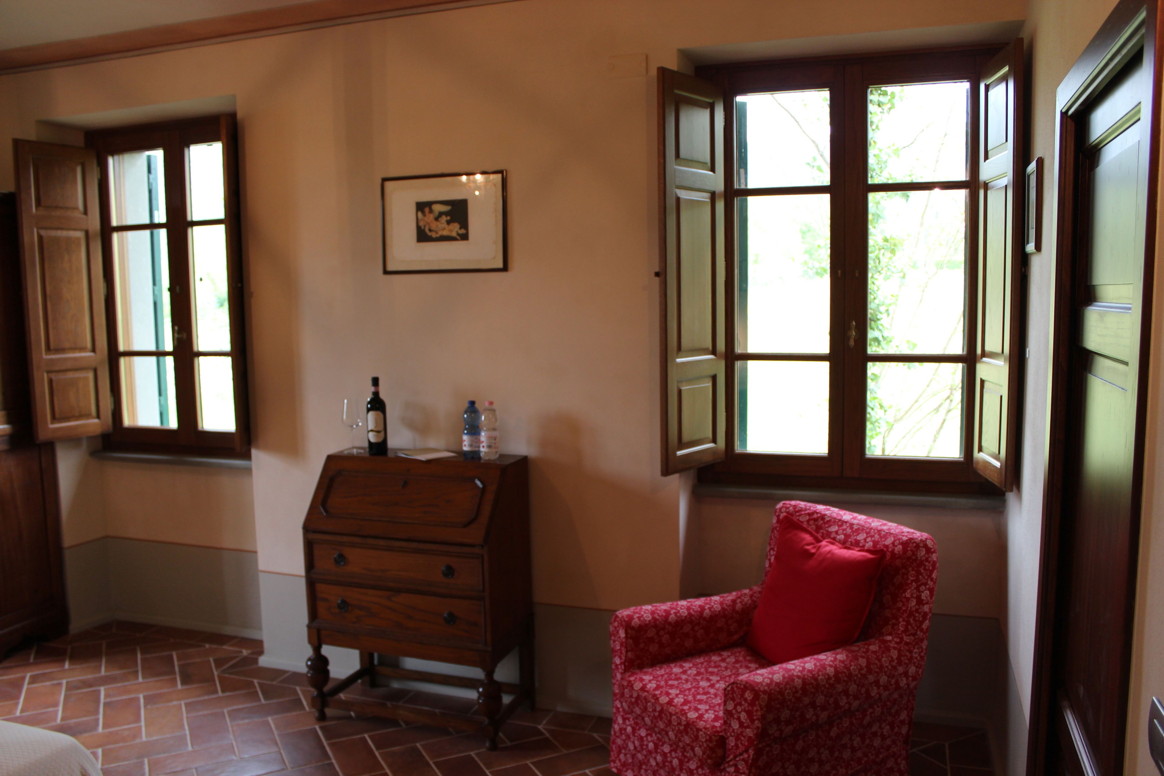 Una habitación con un aparador de madera, un sillón rojo con estampado floral y dos ventanas con contraventanas abiertas.