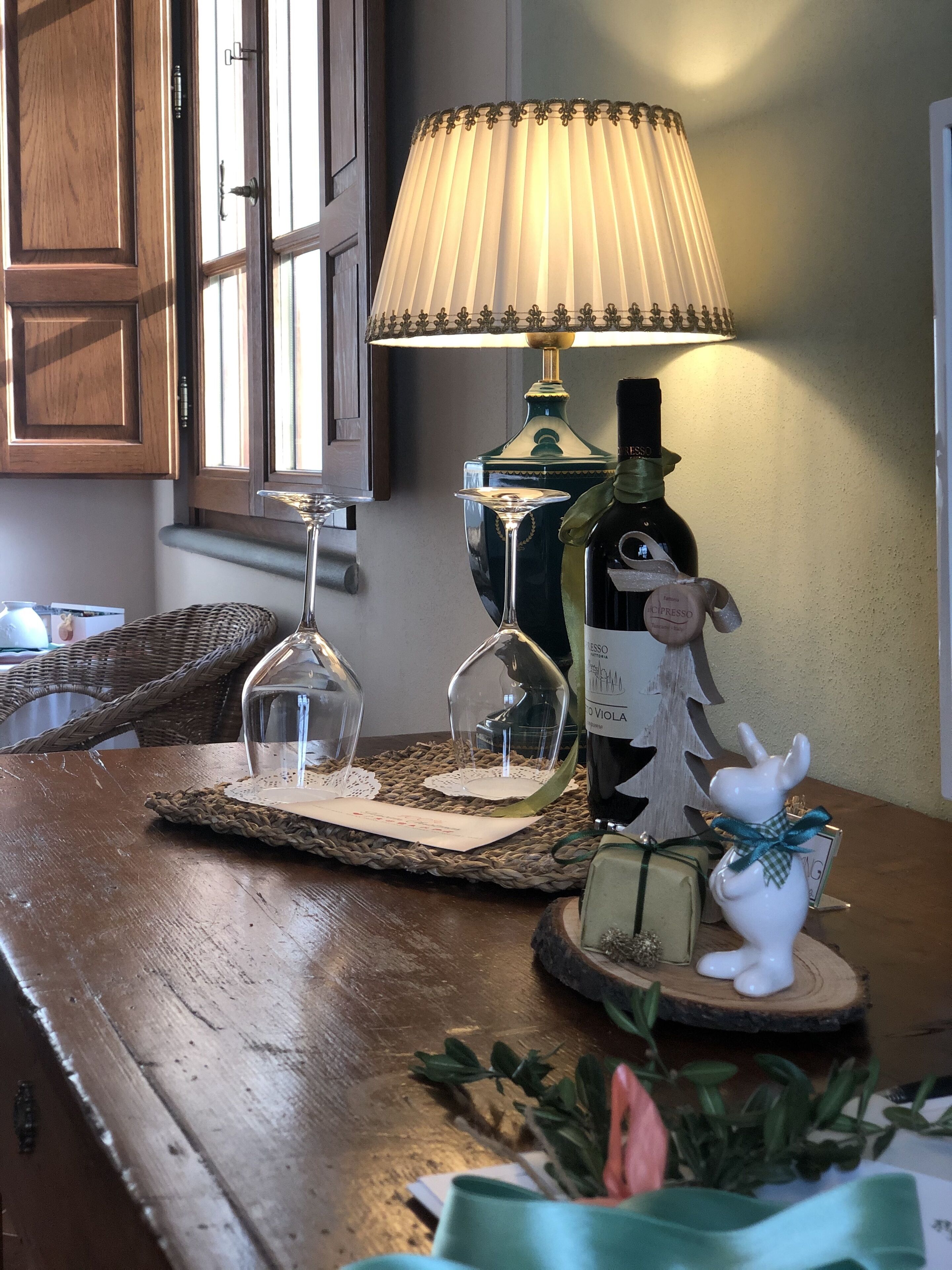 Uma mesa de madeira com uma lâmpada, uma garrafa de vinho, um copo e uma pequena estatueta de cerâmica.