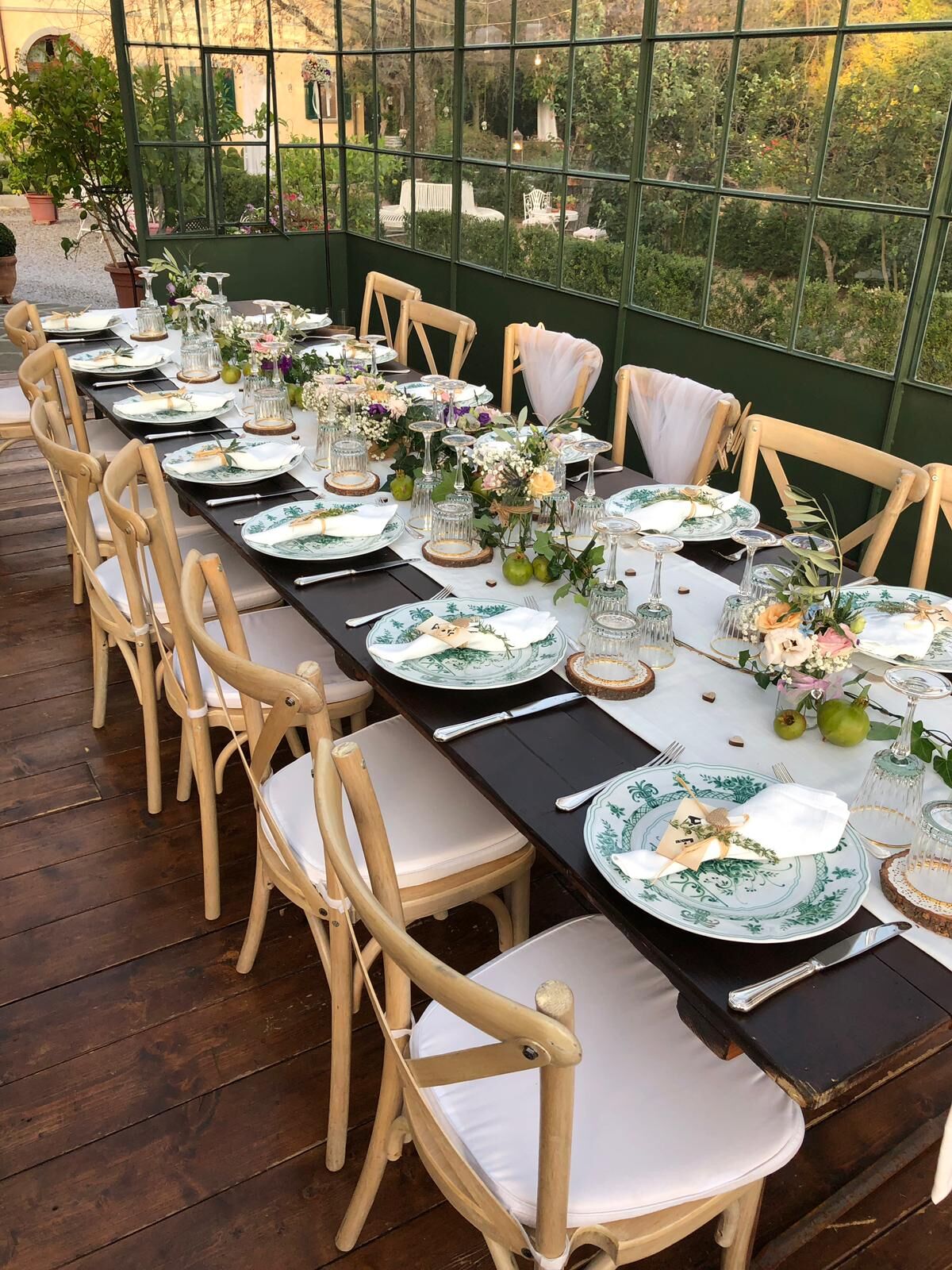Una larga mesa de comedor dispuesta para una comida, con platos blancos y verdes, sillas de madera y centros de mesa florales.