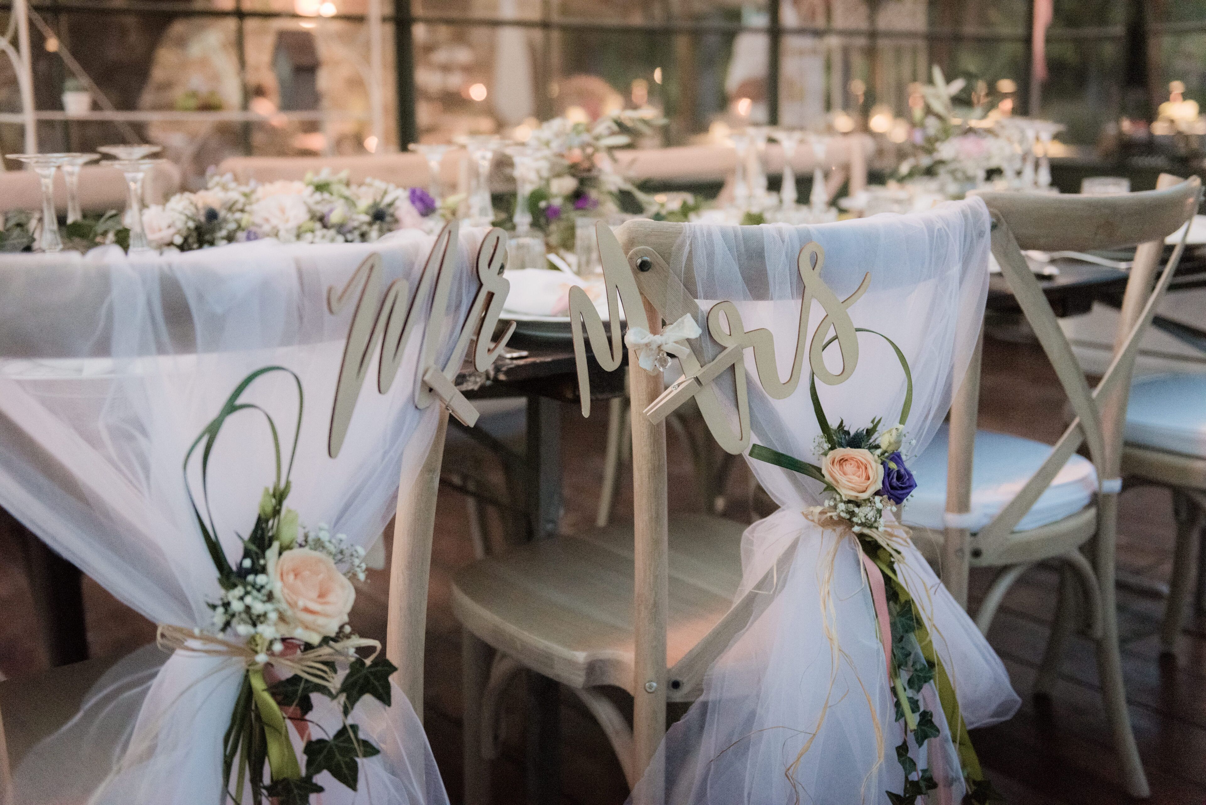 Une réception de mariage avec des tables rondes recouvertes de nappes blanches, des chaises ornées de panneaux indiquant « Monsieur et Madame », et des centres de table floraux.