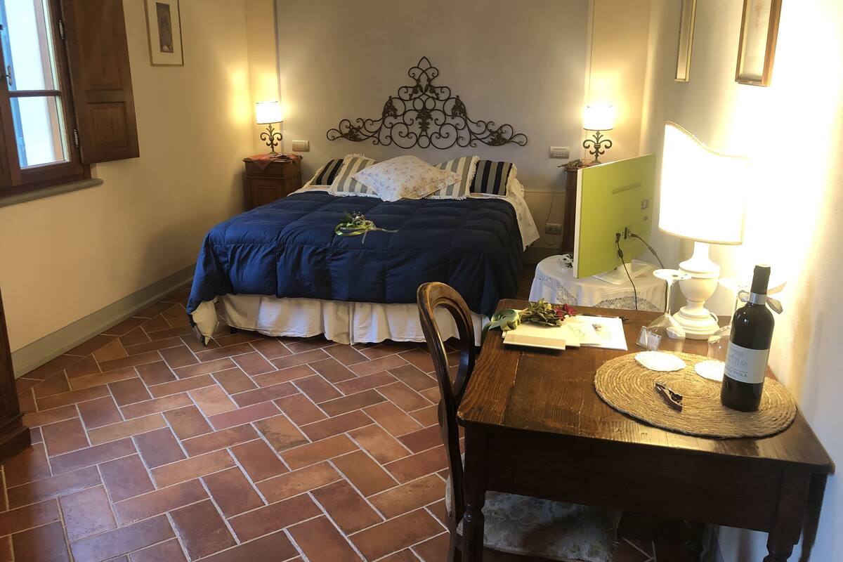 A bedroom with a bed, a bedside table, a desk with a chair, a lamp, a bottle of wine, and a computer on a desk.