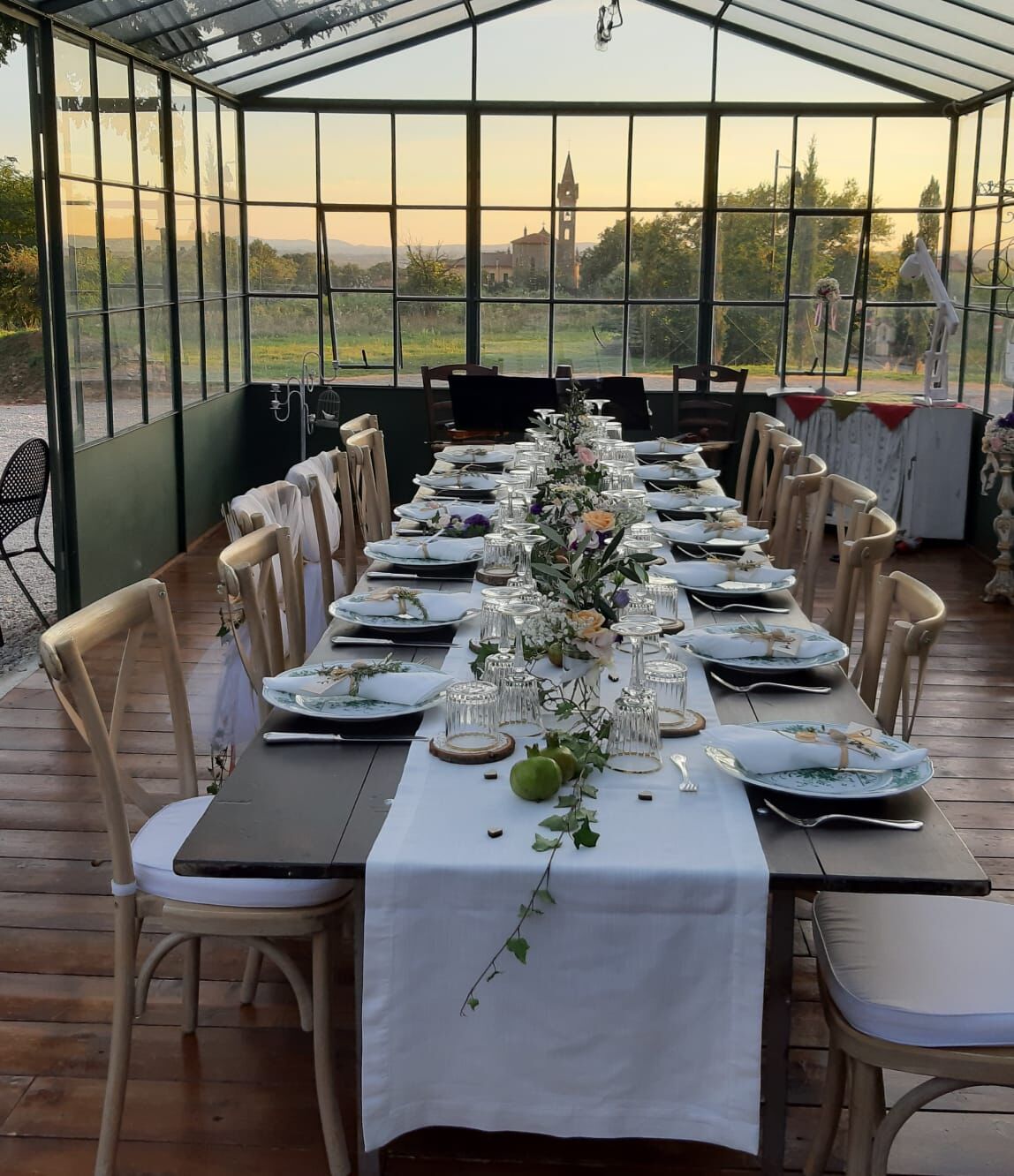 A long table set for a formal outdoor dining event in a greenhouse with a view of a distant building.