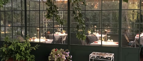 A greenhouse-style dining area with tables and chairs, surrounded by greenery and string lights.