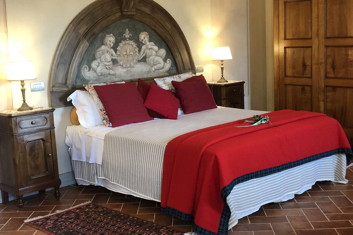 A neatly made bed with a red blanket, two red pillows, and a white bedspread. There are two bedside tables with lamps, a wooden wardrobe, and a patterned rug on the floor.