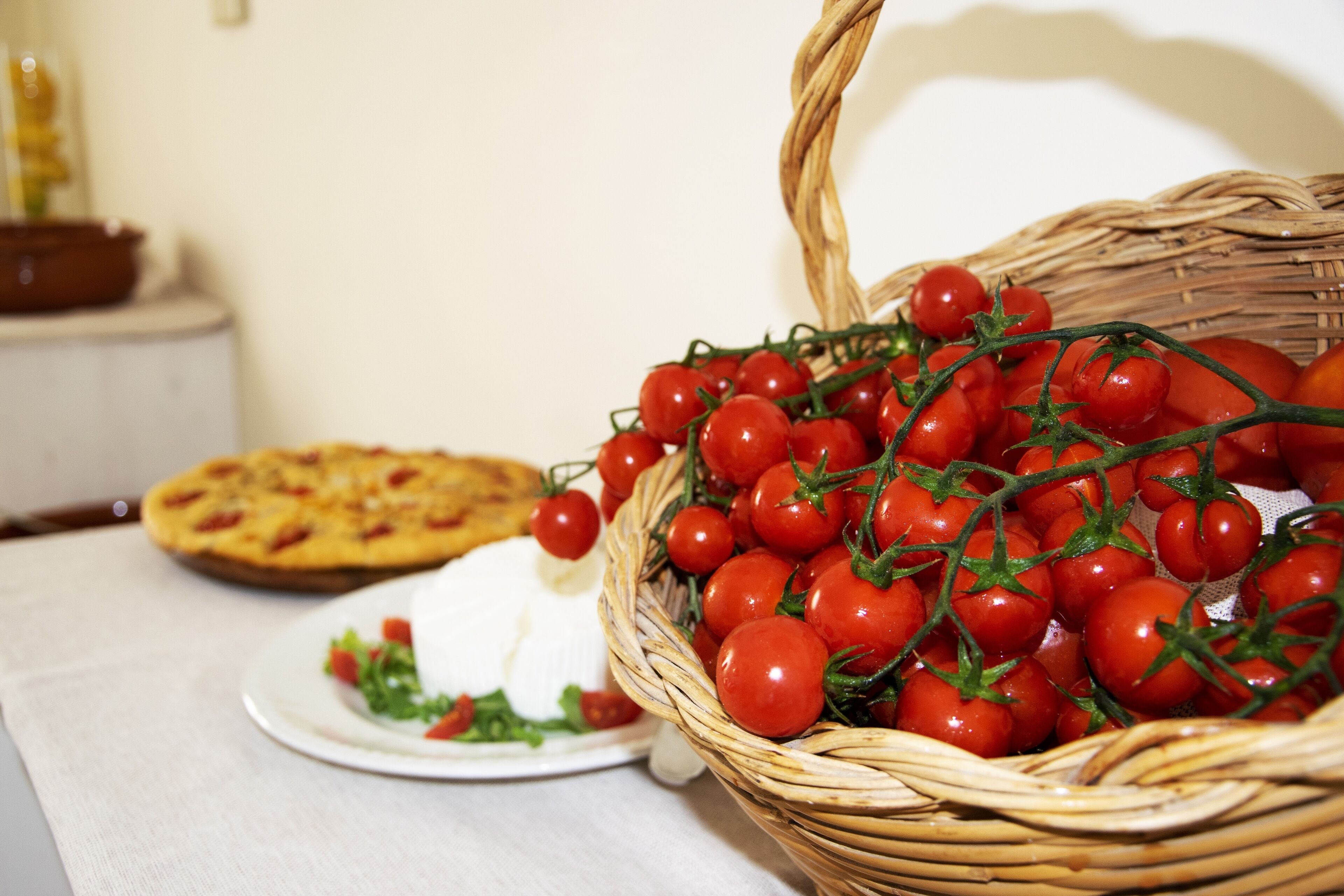 Un panier en osier rempli de tomates cerises est posé sur une table, à côté d’une assiette de salade, et on aperçoit une pizza à l’arrière-plan.