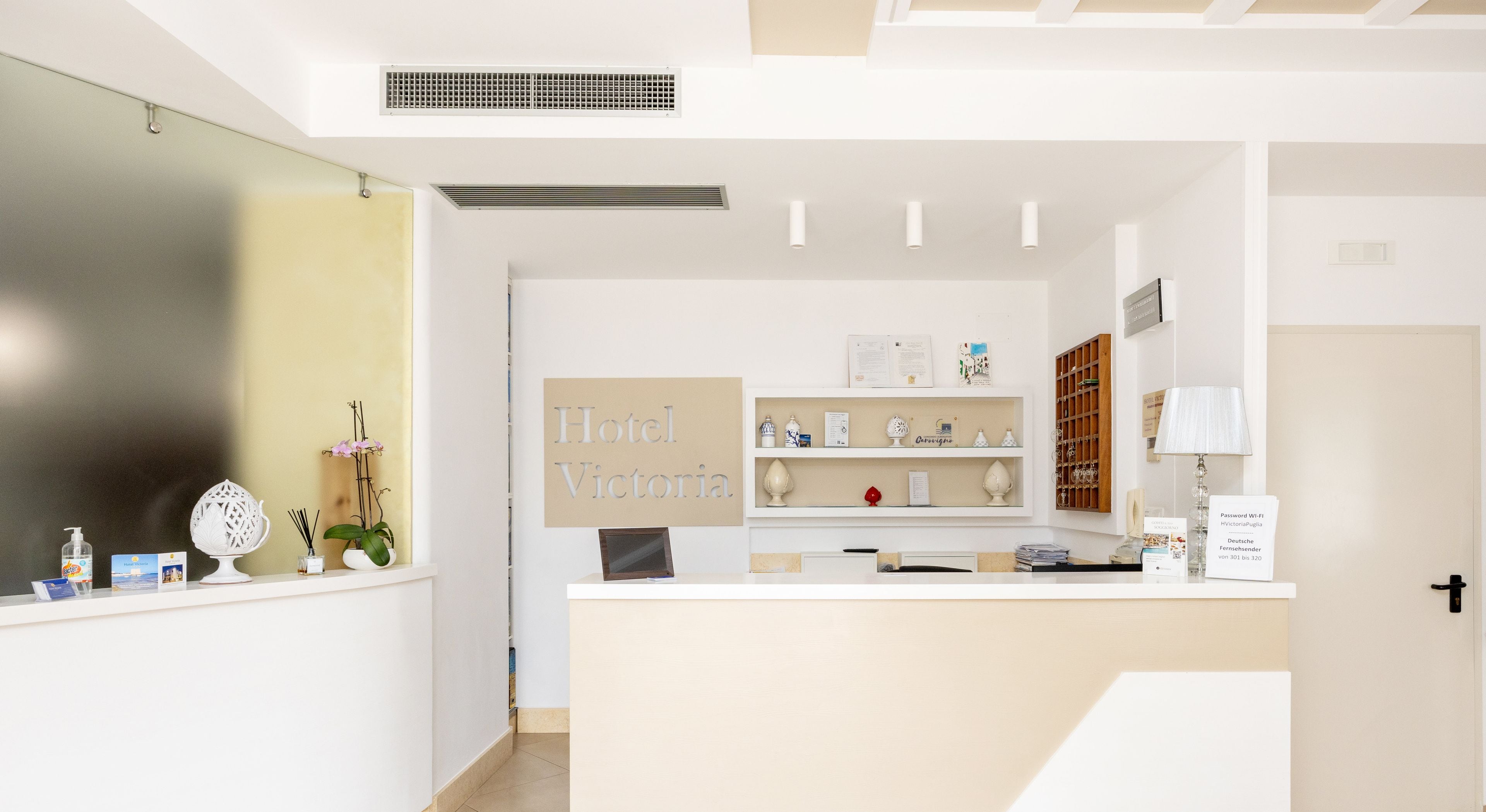 Un espace d’accueil d’hôtel avec un comptoir blanc, un bureau de réception et une enseigne fixée au mur sur laquelle on peut lire « Hôtel Victoria ».