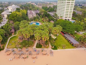 Vue aérienne d’un complexe hôtelier doté d’une piscine, de palmiers et d’une plage aménagée avec des transats et des parasols.