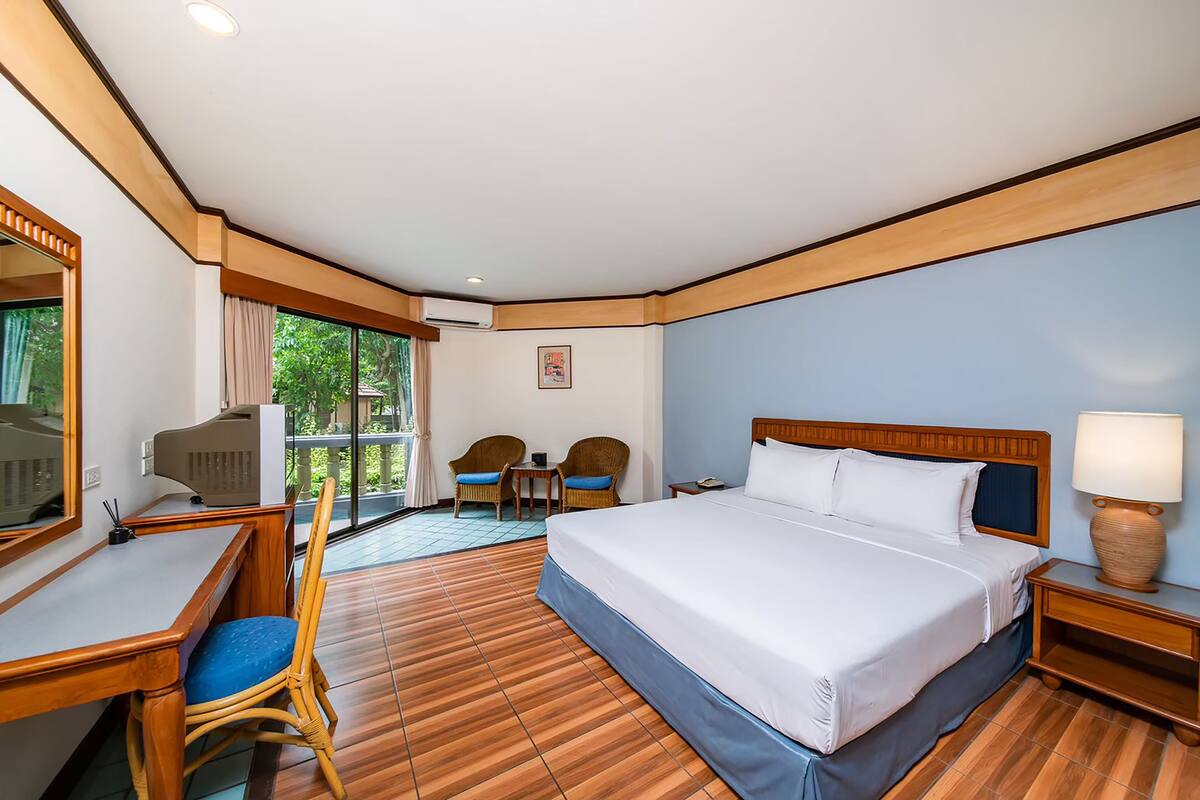 Habitación de hotel con una cama grande, un escritorio, dos sillas, una lámpara y vistas a zonas verdes por la ventana.