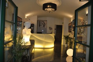 A hotel reception area with a modern design, featuring a curved reception desk, a chandelier, and potted plants.