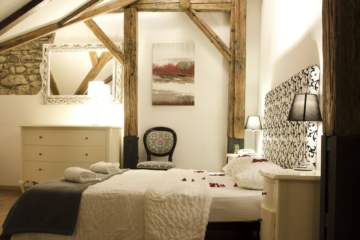 A bedroom with a wooden ceiling, a bed, a nightstand, a chair, and a wall-mounted lamp.