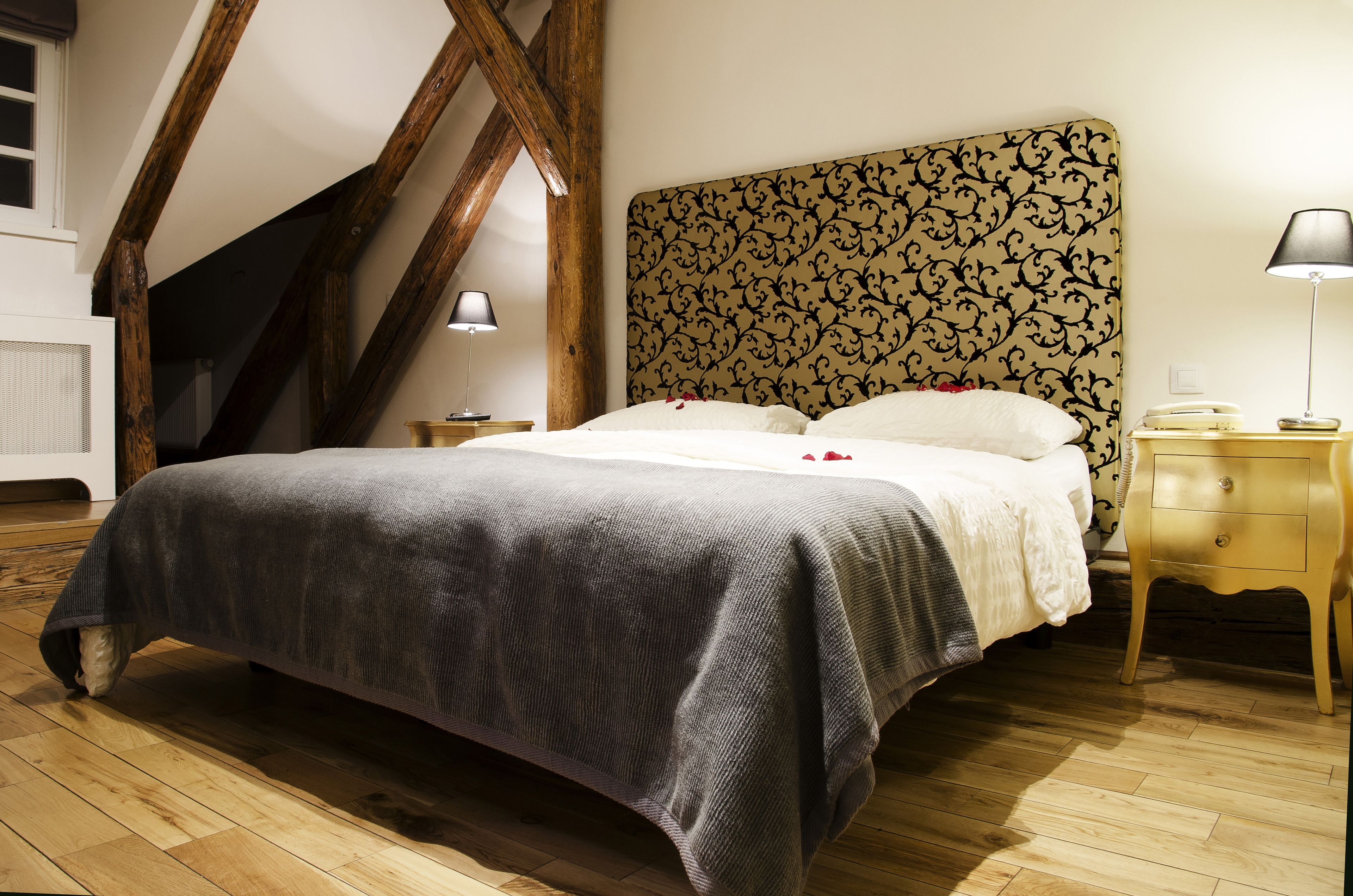 Um quarto com uma cama grande, vigas de madeira, cabeceira estampada, abajures e uma mesinha de cabeceira.