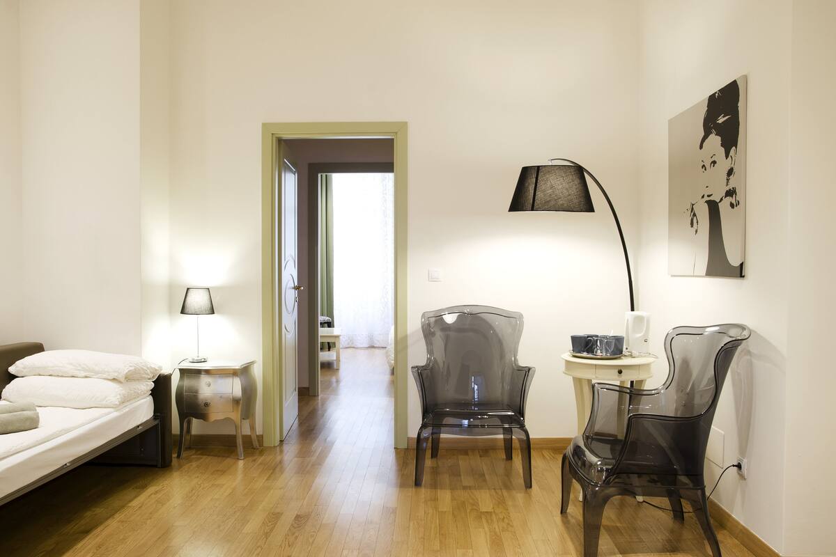 A hotel room with a bed, a desk with a lamp, two chairs, a small table, and a painting on the wall.