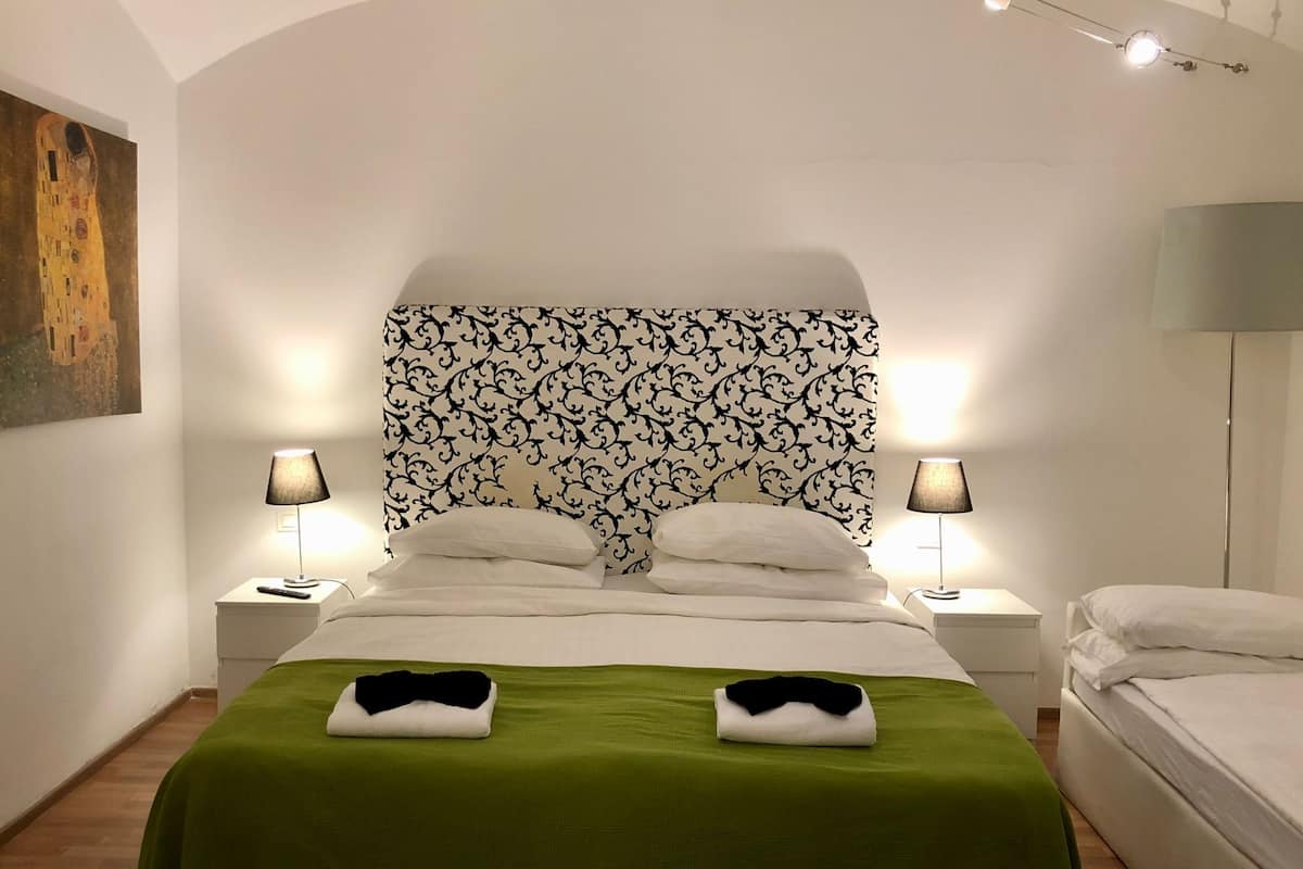 A neatly made bed with a green bedspread, two white pillows, and a black and white patterned headboard. There are two bedside lamps and a painting on the wall.