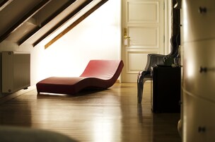 A sunlit room with a red lounge chair, a wooden floor, and a white door.