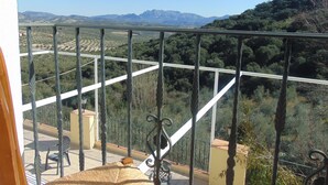 Een balkon met uitzicht op een bergachtig landschap, een witte balustrade en een rieten stoel.