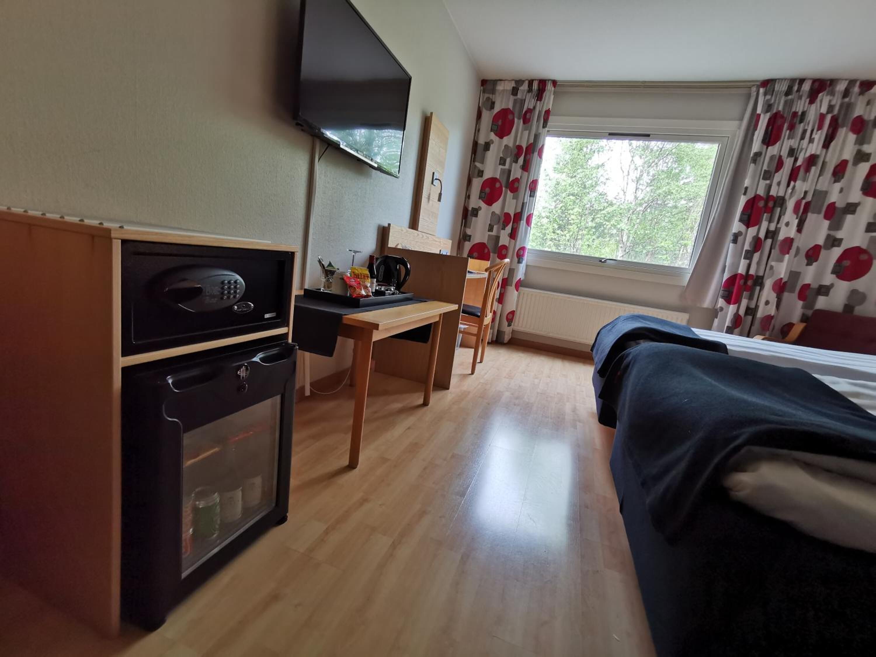 Een hotelkamer met een bed, een bureau met stoel, een magnetron, een kleine koelkast, een televisie en een raam met gordijnen.