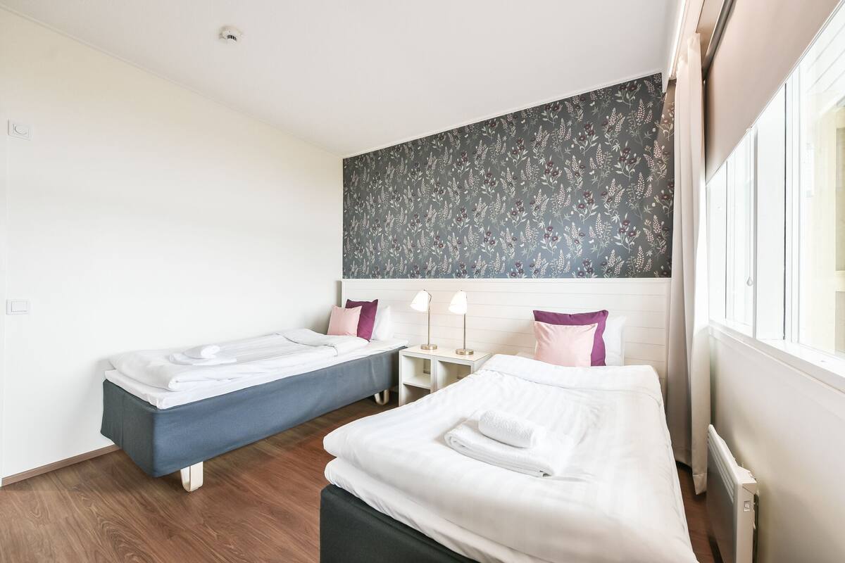 Habitación de hotel con dos camas individuales, una mesita de noche con lámparas, un ventanal grande y un papel pintado con motivos florales.