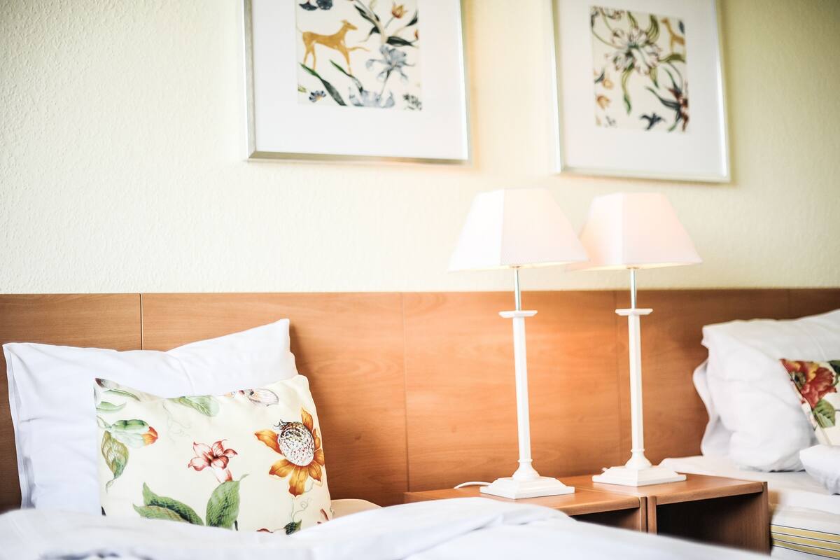 Una cama bien hecha con almohadones florales, un cabecero de madera y una mesita de noche con una lámpara blanca.
