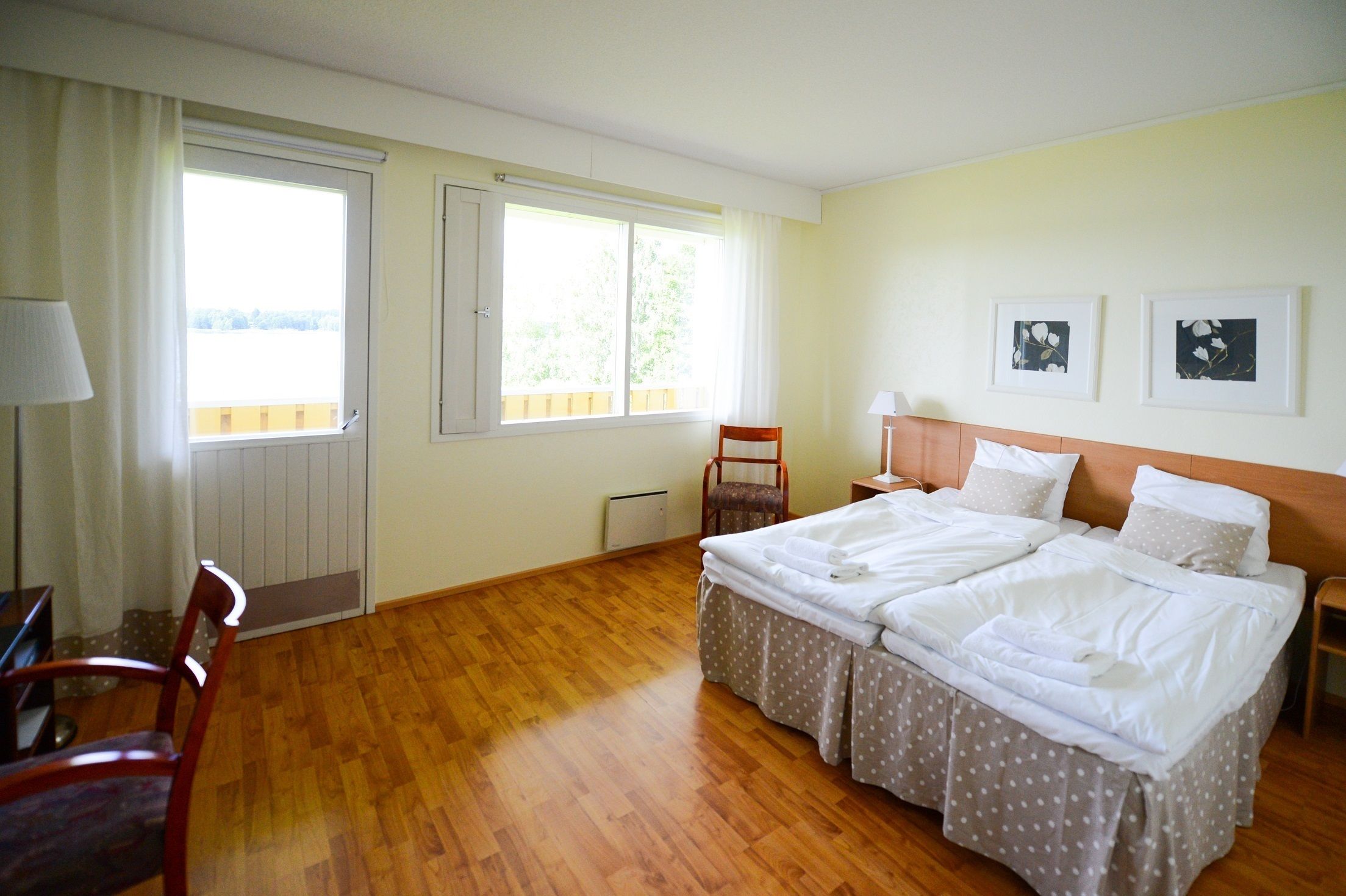 A room with a wooden floor, a bed with white bedding, a chair, a desk, and a window with curtains.