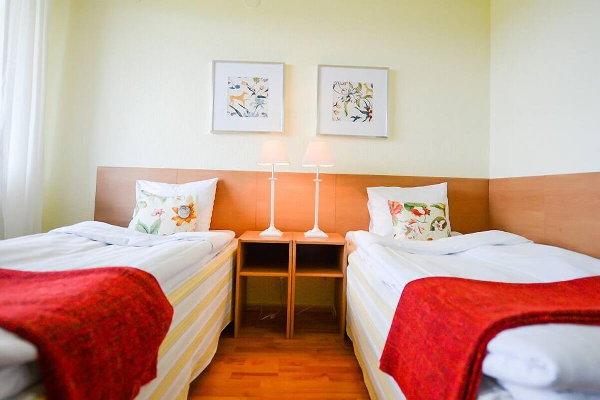 Habitación de hotel con dos camas individuales, cada una con una manta roja y almohadas estampadas, cabeceros de madera y dos lámparas fijadas a la pared.