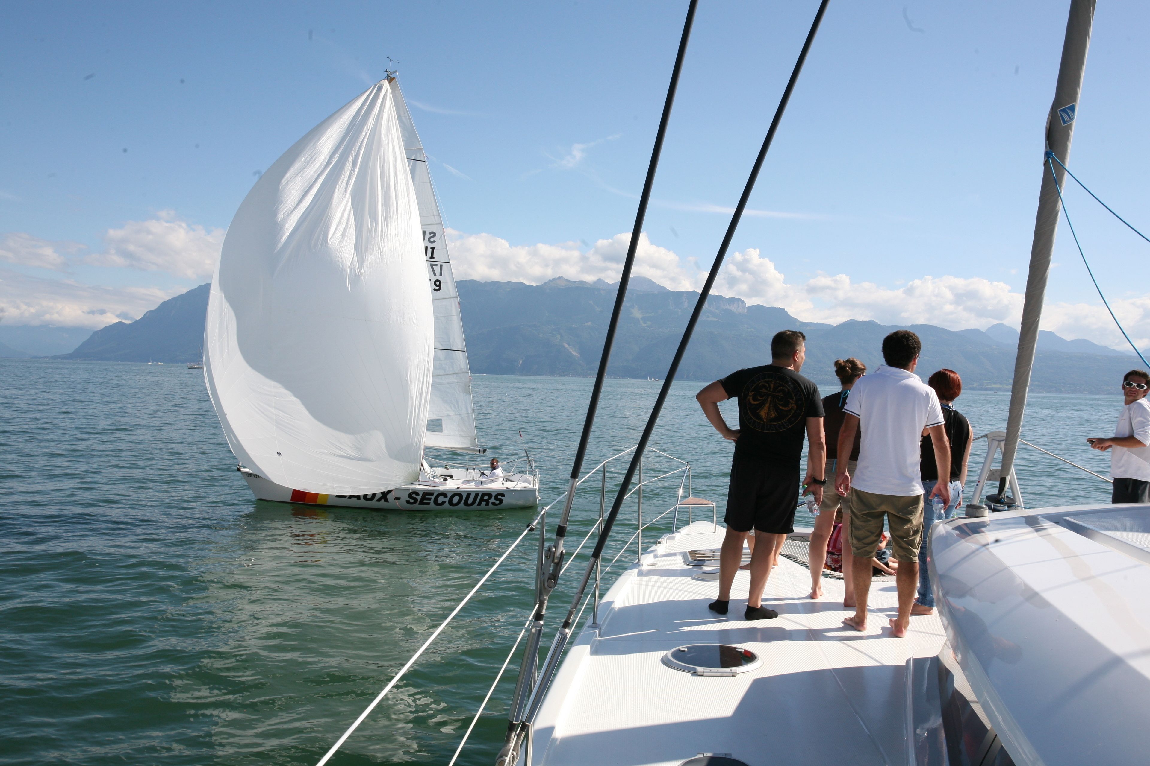 Ein Segelboot mit weißer Segel namens "RUI - SECOURS" auf ruhigem Wasser, mit einer Bergkulisse im Hintergrund.
