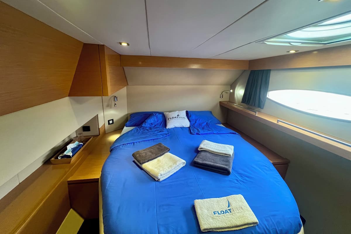 Ein kleines, modernes Schlafzimmer mit blauer Bettdecke, ordentlich gefalteten Handtüchern und einem Fenster mit Tageslicht.