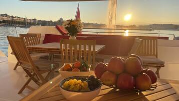Una mesa con frutas y un florero en la cubierta de un barco, con vista al agua y edificios.