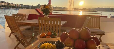 A table with fruits and a vase of flowers on a boat deck with a view of the water and buildings.