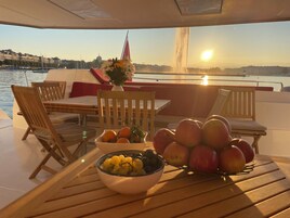 Una mesa con frutas y un florero en la cubierta de un barco, con vista al agua y edificios.