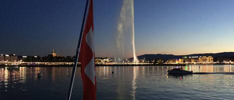A nighttime view from a boat with a flag, a fountain, and city lights in the background.