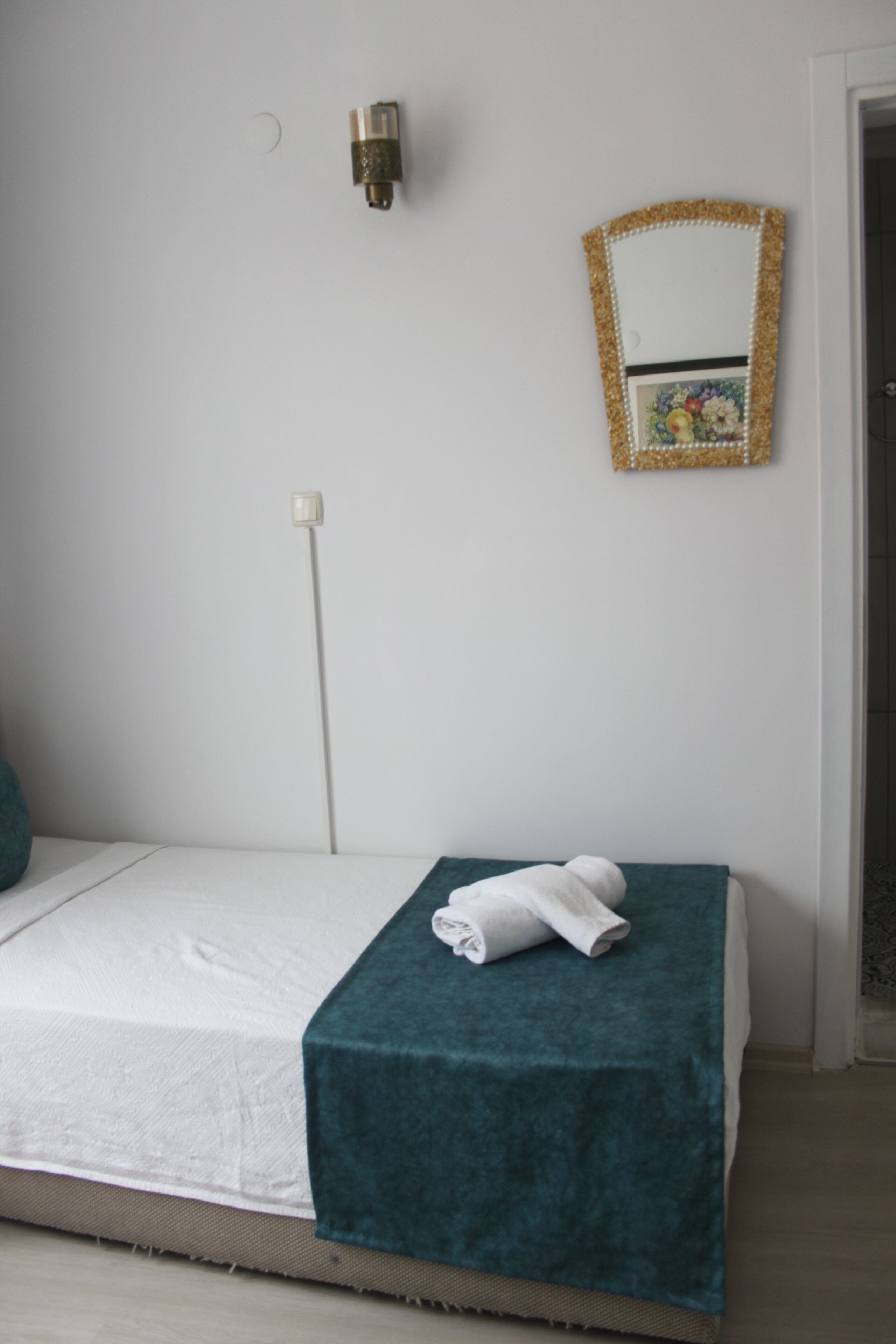 A neatly made bed with a teal blanket and a folded white towel, a wall-mounted mirror with a decorative frame, and a plain white wall with a single light switch.