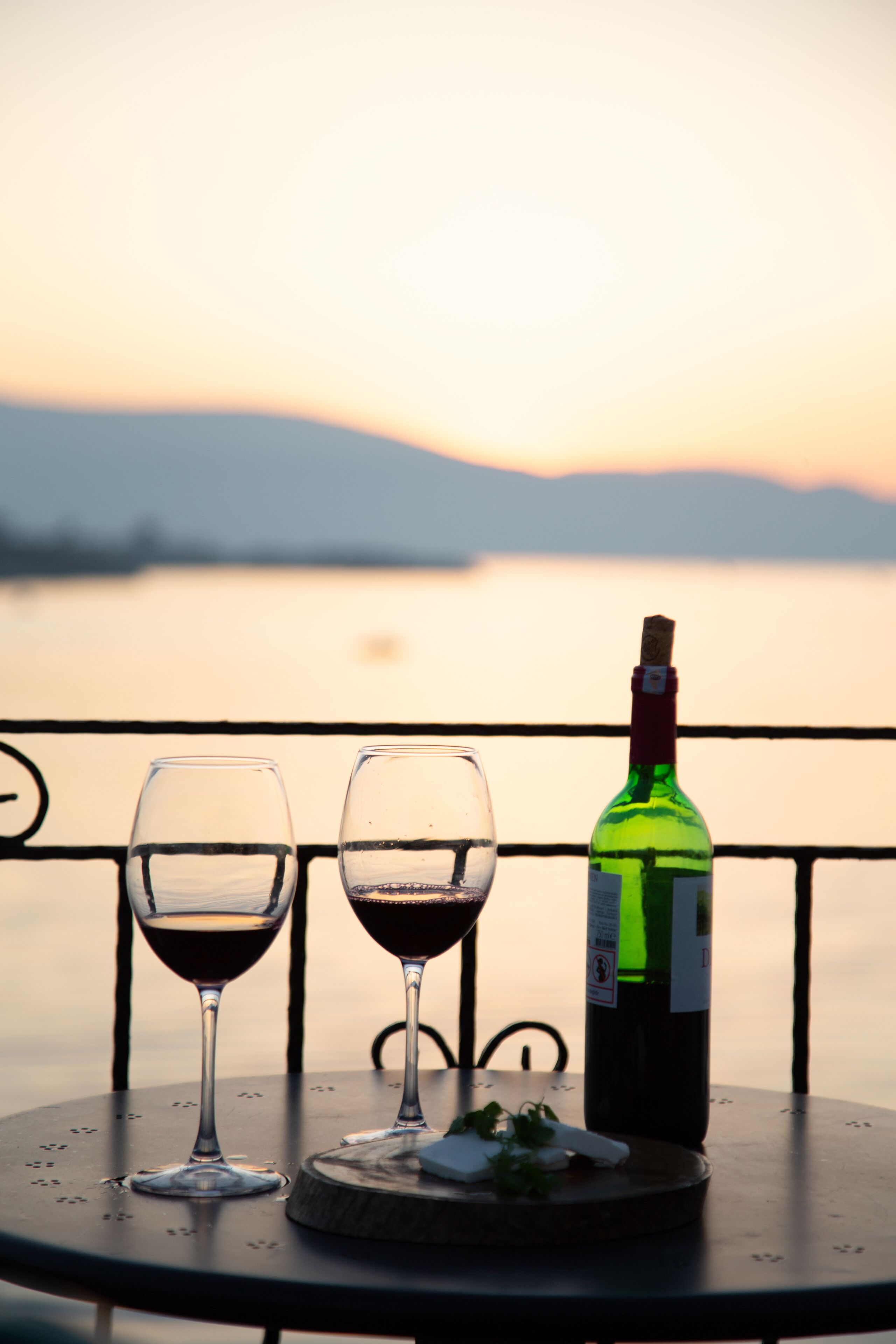 Una mesa con dos copas de vino y una botella, frente a un paisaje con el atardecer sobre el agua.