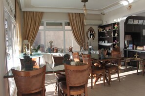 A dining area with a glass table, wooden chairs, and a bookshelf with decorative items. - Foca Kumsal Hotel (Foça)
