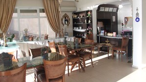 A dining area with a glass table, wooden chairs, and a kitchen in the background. - Foca Kumsal Hotel (Foça)