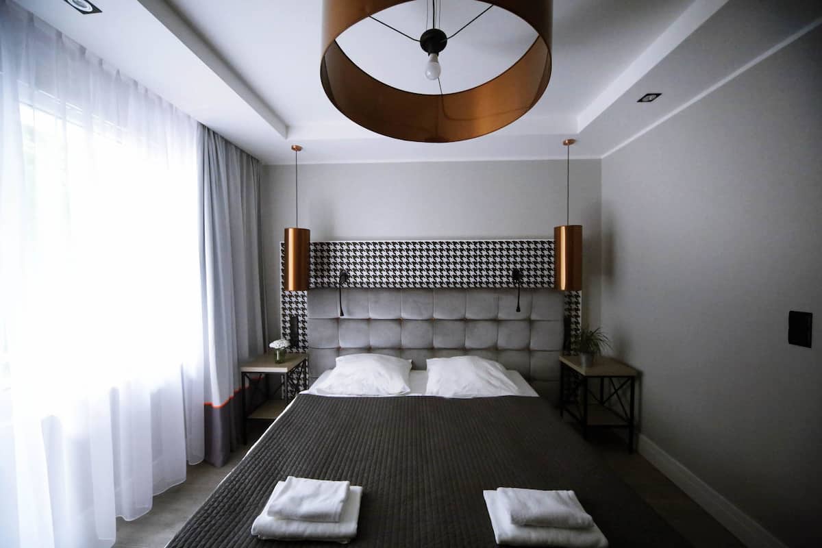 A modern hotel room with a large bed, a tufted headboard, a round ceiling light, and a view of the outdoors.