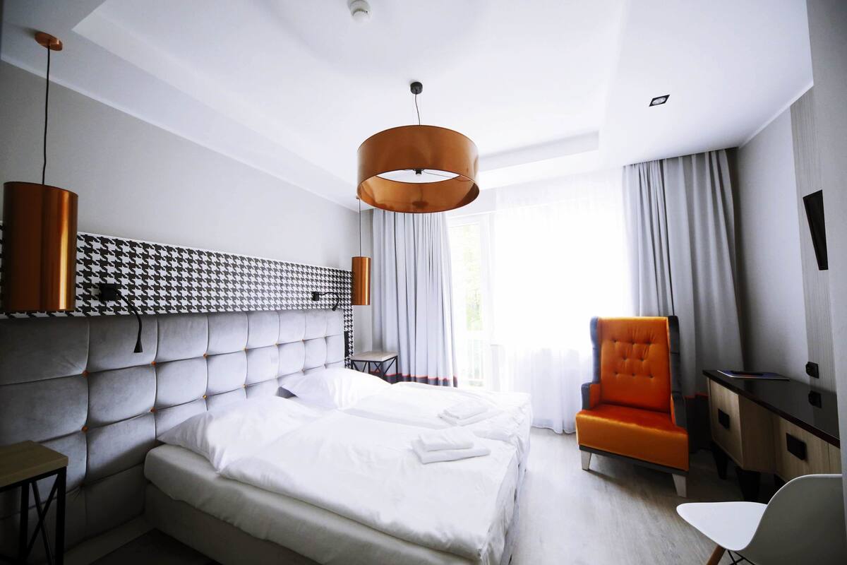 A modern hotel room with a large bed, a desk, an orange armchair, and a white chair.