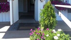 En byggnadsentré med en träveranda, en metalldörr och krukväxter med blommor.