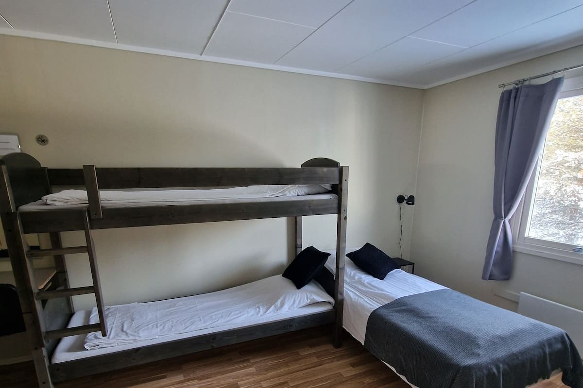A dormitory room with two bunk beds, a single bed, wooden flooring, a ceiling fan, a window with curtains, and a wall-mounted light fixture.