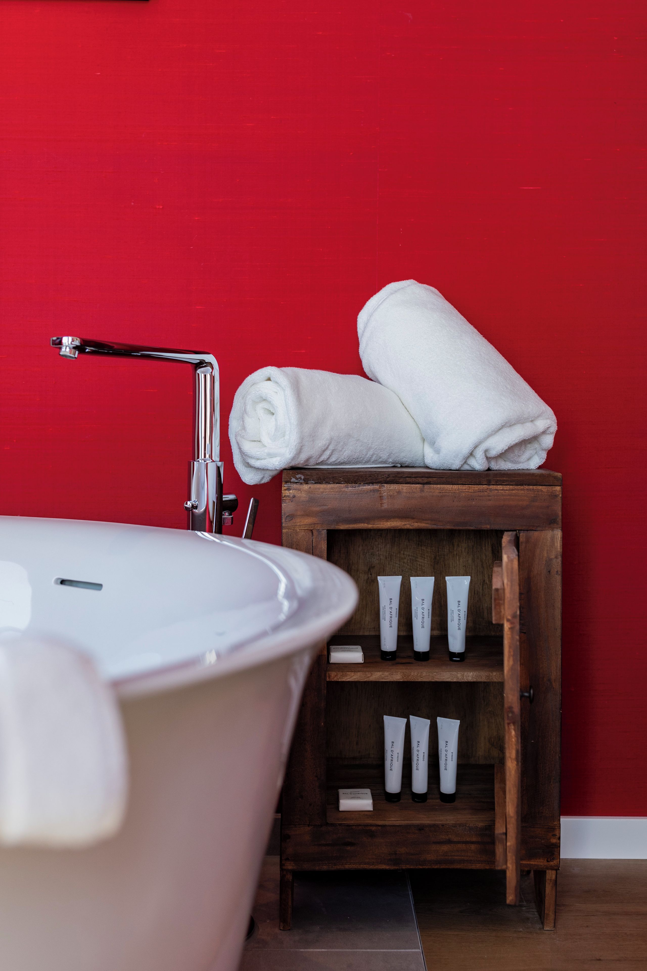 Un baño con una bañera blanca, una pared roja y un estante de madera con artículos de aseo.