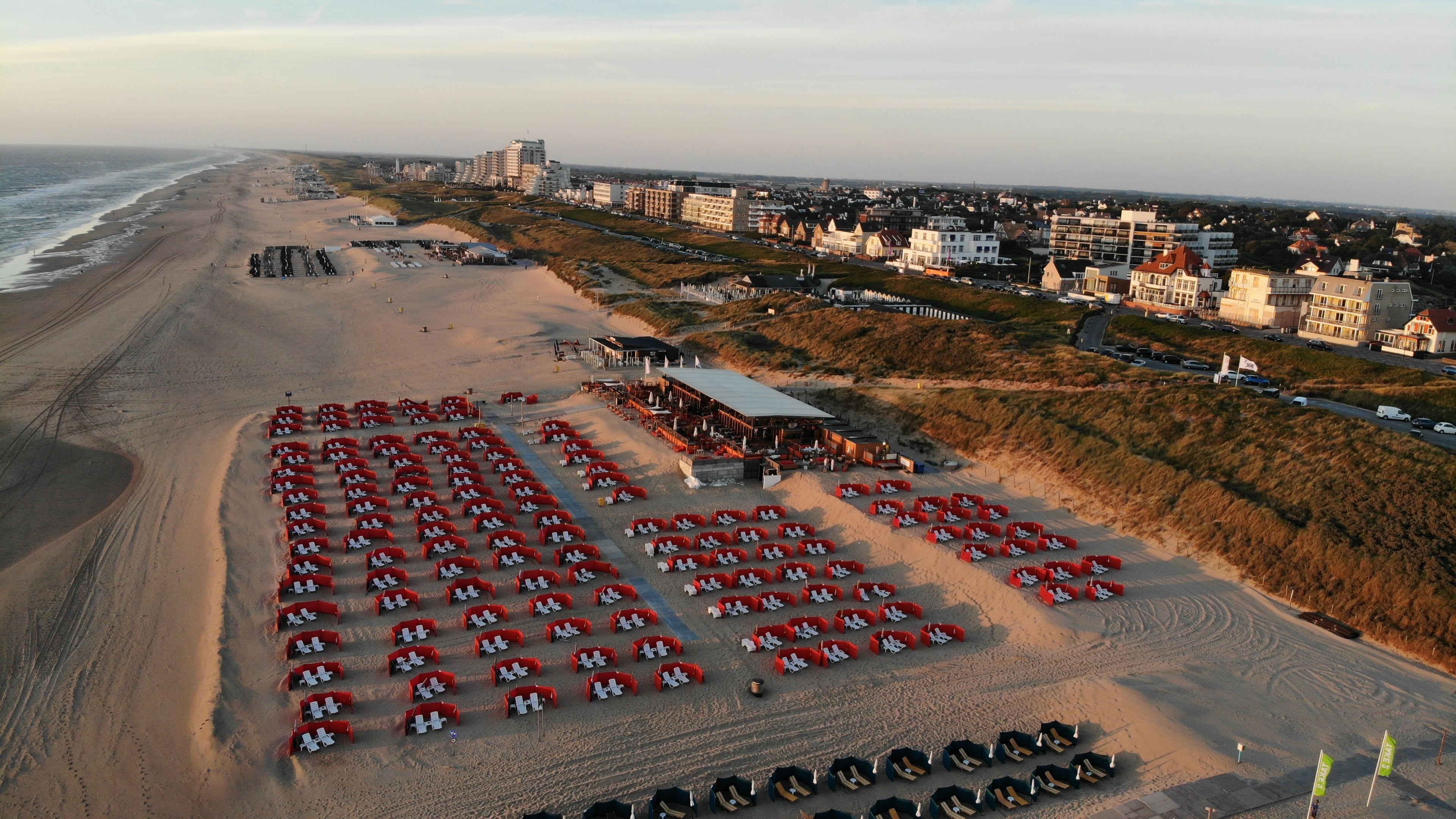 Eine Luftaufnahme eines Hotels direkt am Strand, mit mehreren Reihen von Liegestühlen und Sonnenschirmen, einem großen Gebäude mit flachem Dach und einer Küstenlinie im Hintergrund.