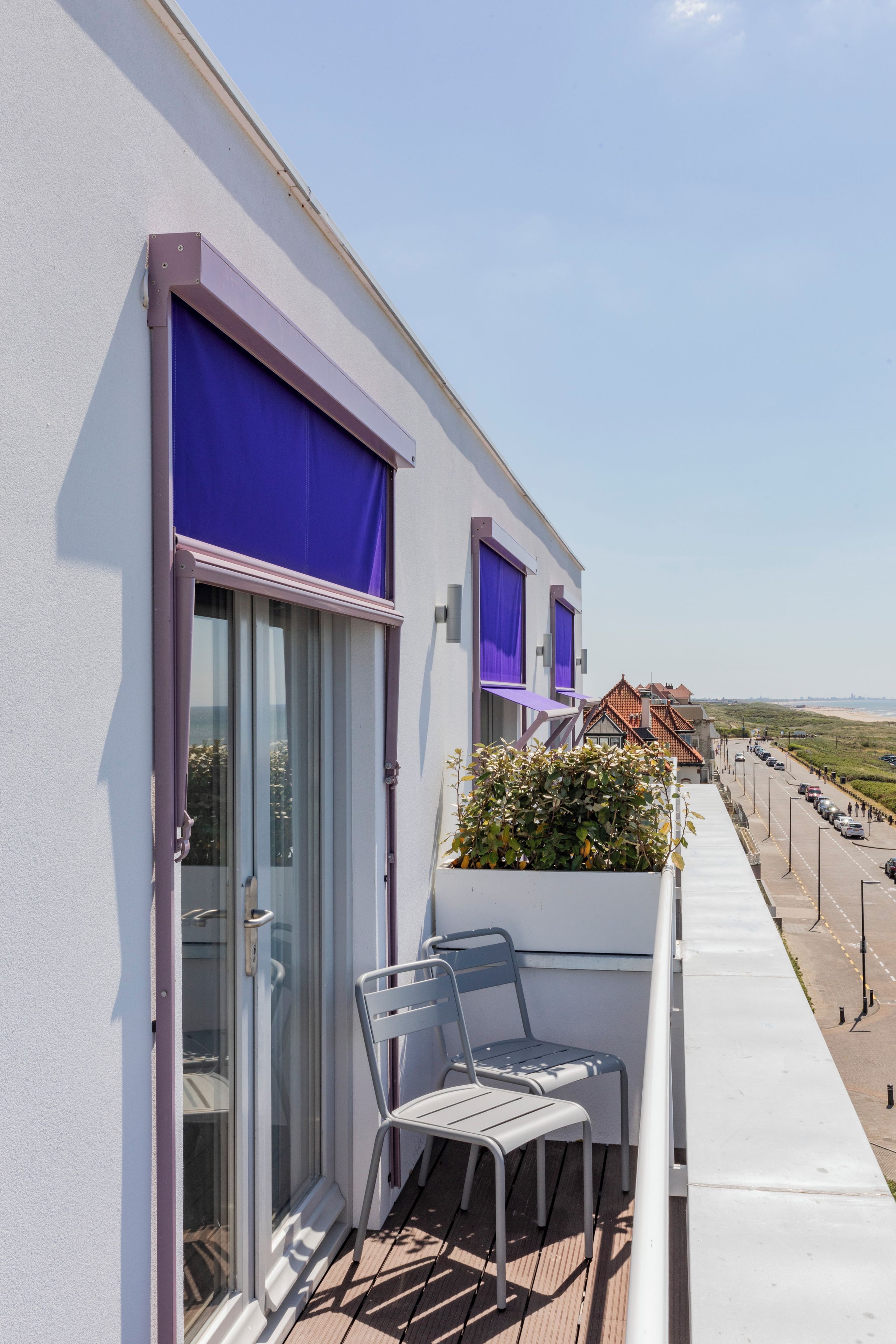 A balcony with two chairs and a small table, a window with a purple blind, and a view of a beach and buildings in the distance.