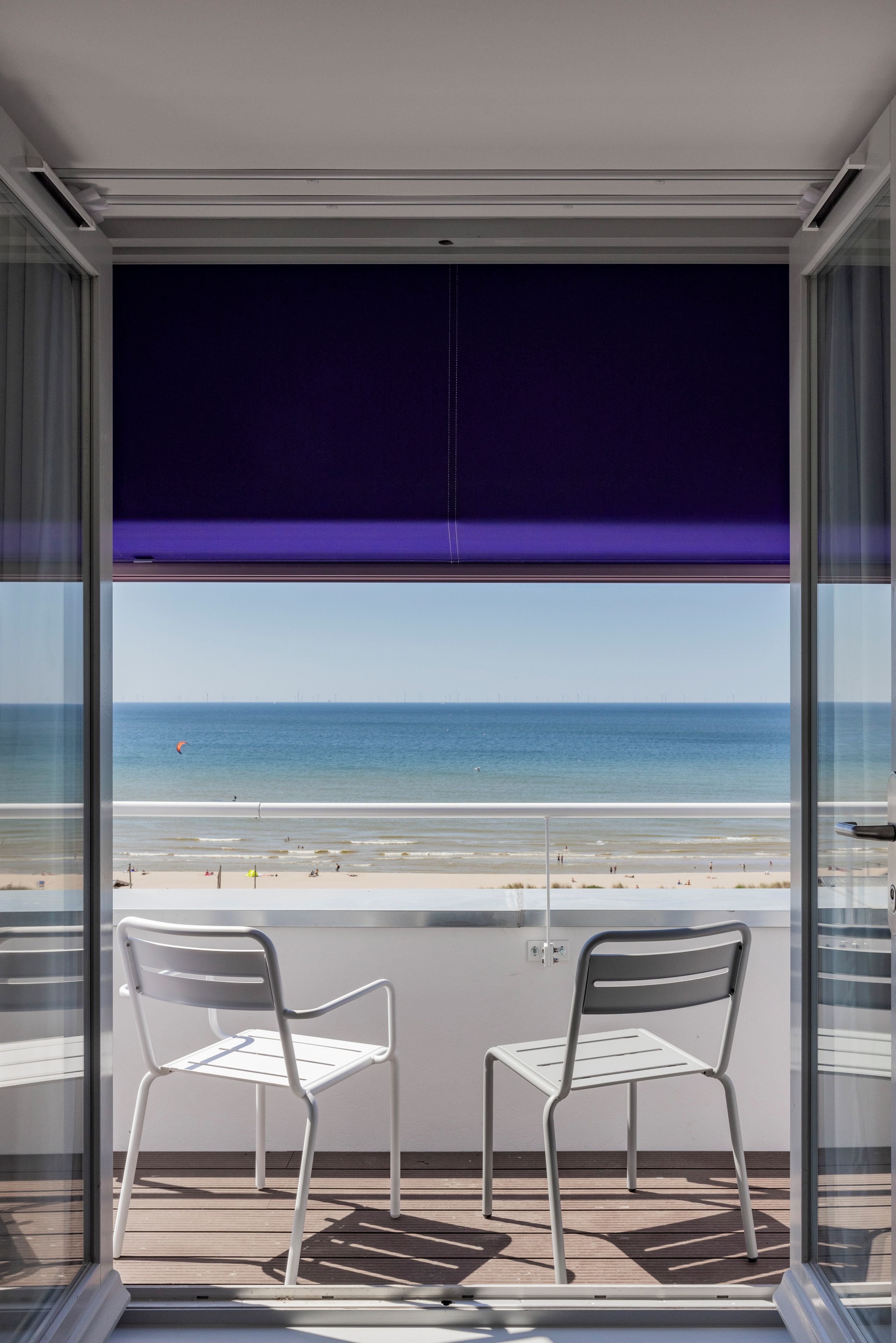 A balcony with two white chairs, a view of the beach, and a window with a purple blind.