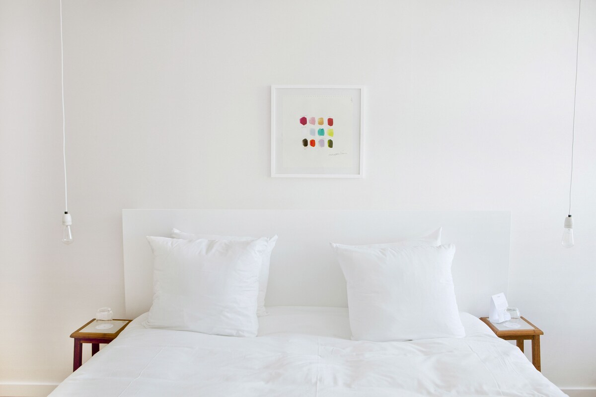 A neatly made bed with white bedding, two bedside tables with a small lamp on each, and a framed artwork on the wall.